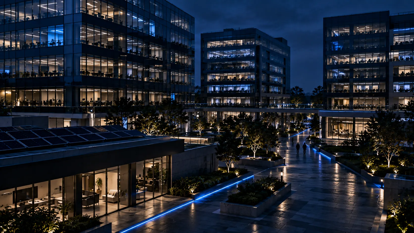 A technology park at night with glass office towers, rooftop solar panels on a lower building and a plaza with a thin blue line of light along the walkway.